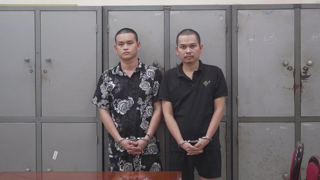 Hai người đàn ông mặc thường phục đứng cạnh nhau, tay bị còng, phía sau là dãy tủ sắt màu xám trong một căn phòng - Ảnh 1.