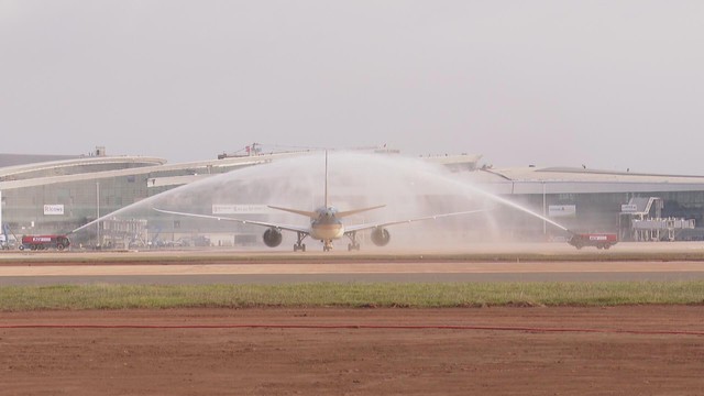 Một chiếc máy bay thương mại cỡ lớn đang lăn bánh trên đường băng sân bay rộng lớn, được chào đón bằng nghi thức phun vòi rồng từ hai chiếc xe cứu hỏa hai bên, phía sau là công trình nhà ga sân bay đang được xây dựng - Ảnh 1.