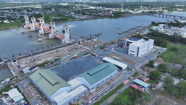 Góc máy trên cao bao quát một khúc sông rộng với hệ thống cống ngăn triều lớn, trên bờ là khu vực công trường đang thi công cùng một số tòa nhà mái xanh và một tòa nhà điều hành màu trắng - Ảnh 1.