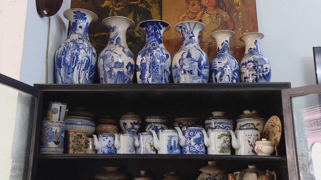Một chiếc tủ gỗ trưng bày nhiều món đồ gốm sứ cổ, trong đó ngăn trên cùng xếp hàng sáu chiếc bình hoa lớn men xanh trắng họa tiết phong cảnh, ngăn dưới là các loại ấm trà, hũ, thố đa dạng kích cỡ và hoa văn - Ảnh 2.